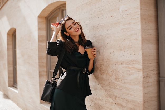 Lifestyle portrait of charming young brunette with bright makeup, dark outfit with dress, jacket, bag, sunglasses and belt standing near beige wall outdoors and smiling