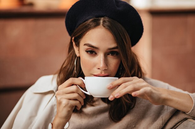 Lifestyle portrait of elegant young woman with dark wavy hairstyle, trendy makeup, fashionable beige pullover and coat, sitting at cafe terrace and drinking coffee from white cup