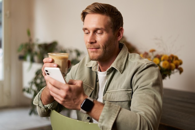 Бесплатное фото Портрет красивого молодого человека, сидящего в кафе, проверяющего свой телефон и пьющего кофе.