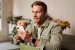 Бесплатное фото Портрет красивого молодого человека, сидящего в кафе, проверяющего свой телефон и пьющего кофе.
