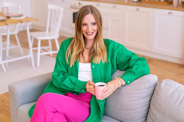 Lifestyle portrait of stylish blonde woman relaxed on her sofa, wearing green linen shirt and fucsia pants, drinking morning coffee.