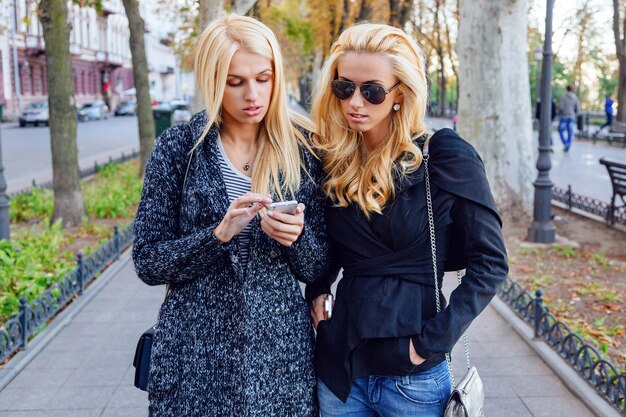 Lifestyle portrait of two best friends blonde girls spending time in the center of the city at nice fall autumn day, using smartphone, wearing sunglasses ant trendy fashion looks.