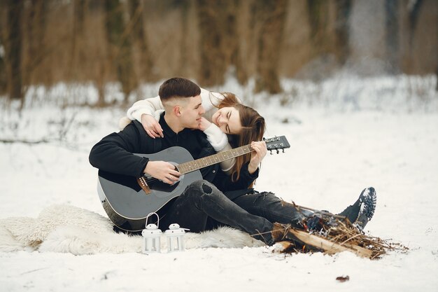 눈 덮인 숲에 앉아 부부의 라이프 스타일 샷. 겨울 휴가를 야외에서 보내는 사람들. 커플은 기타를 재치.