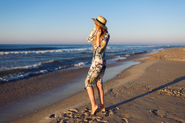 Lifestyle summer fashion portrait of beauty blonde woman posing at lonely beach, wearing bikini stylish pareo and hat, look at ocean, luxury vacation mood, bright toned colors.
