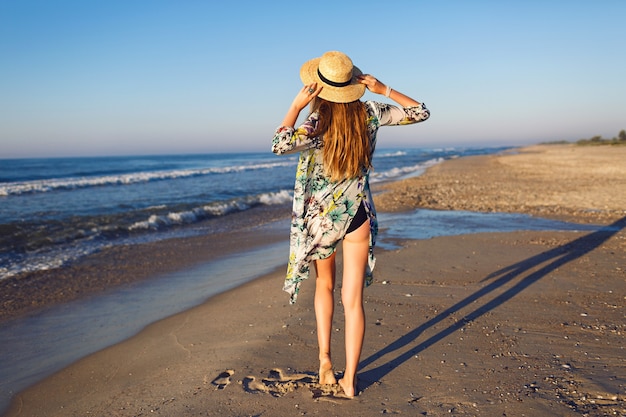 Lifestyle summer fashion portrait of beauty blonde woman posing at lonely beach, wearing bikini stylish pareo and hat, look at ocean, luxury vacation mood, bright toned colors.