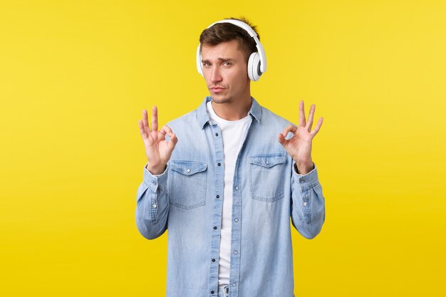 Lifestyle, summer holidays, technology concept. Cool pleased handsome guy in casual clothes, listening music in headphones, showing okay gesture, like awesome earphones, yellow background.