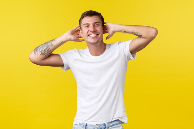 Lifestyle, summer and people emotions concept. Carefree handsome happy man with white cheerful smile, holding hands behind head, enjoying vacation, standing yellow background.