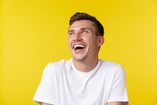 Lifestyle, summer and people emotions concept. Close-up portrait of carefree happy handsome man, looking upper left corner banner and laughing, standing in basic white t-shrit over yellow background.