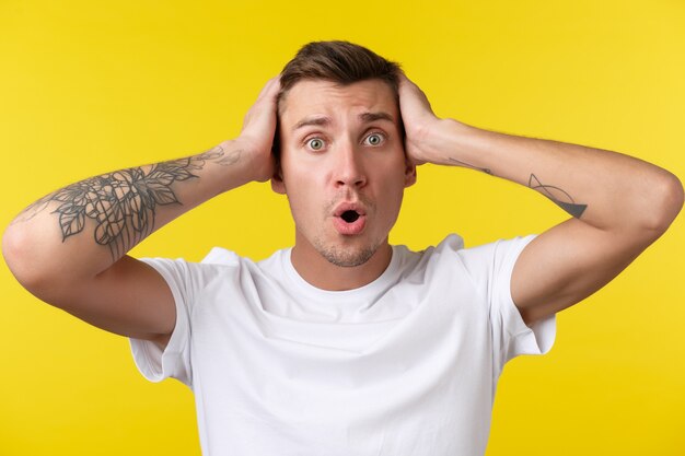 Lifestyle, summer and people emotions concept. Close-up portrait of worried and nervous young man grab head in denial, having huge trouble, panicking, feeling anxious over yellow background.