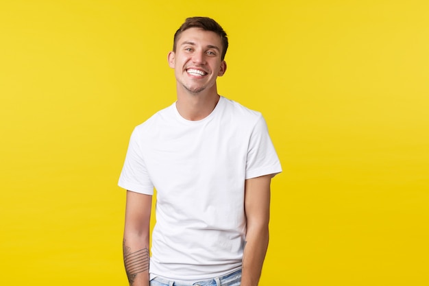 Концепция образа жизни, лета и людей эмоции. Красивый харизматичный кавказский парень в повседневной белой футболке, широко улыбаясь идеальной белой улыбкой, стоит на радостном желтом фоне.