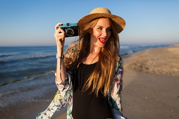 Free photo lifestyle sunny portrait of young beauty photographer woman posing near lonely beach in the front of the ocean, stylish bikini hat sunglasses and pareo, luxury vacation vibes.