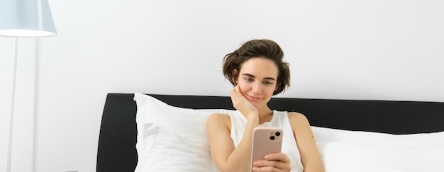 Free photo lifestyle and technology concept smiling beautiful woman lying in bed with smartphone resting in