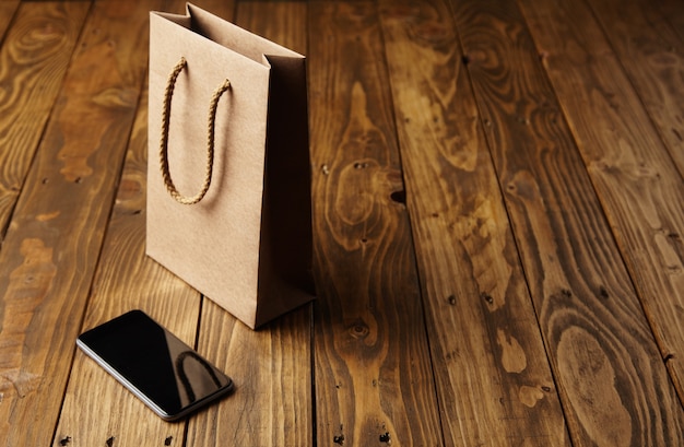 Светло-коричневый бумажный пакет, отражающийся в безупречном черном смартфоне, лежащем рядом с ним на деревянном столе ручной работы.