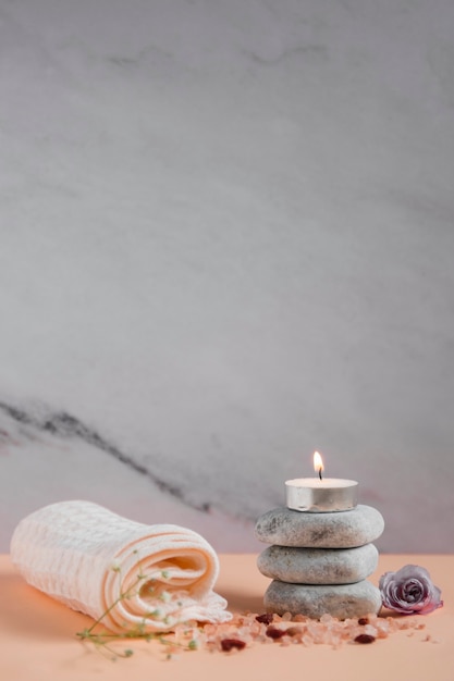 Free photo lighted candle over the spa stones with napkin; rose and himalayan salts on peach colored backdrop against grey background