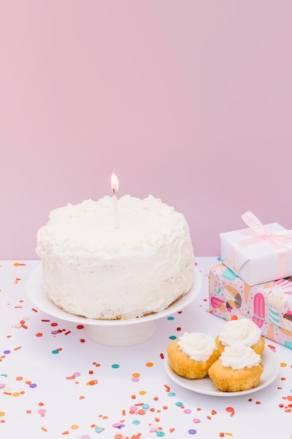 Зажженная свеча на белом торте с кексами и подарками на белом столе на розовой стене