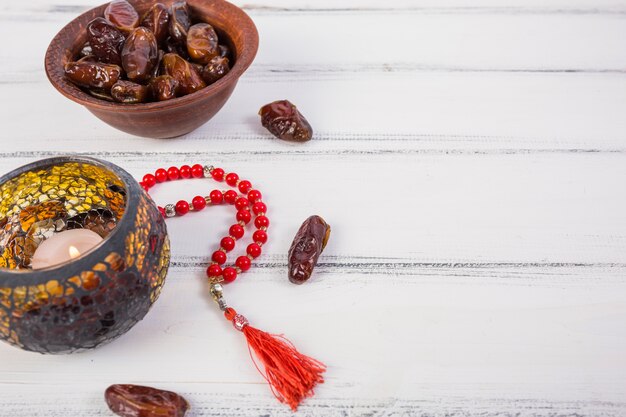 Lighted candle with bowl of juicy dates and red prayer beads on white wooden background