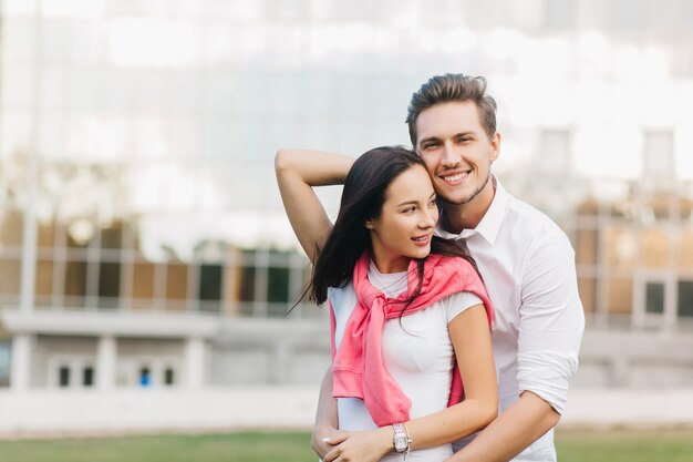 Lightly-tanned brunette woman looking away while smiling boyfriend embracing her