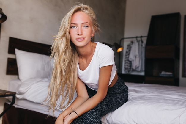 Lightly-tanned cute woman with long hairstyle sitting on bed. woman posing in morning.