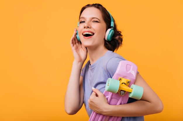 Lightly-tanned merry girl posing with pink longboard. Indoor photo of laughing cute lady in big headphones isolated.