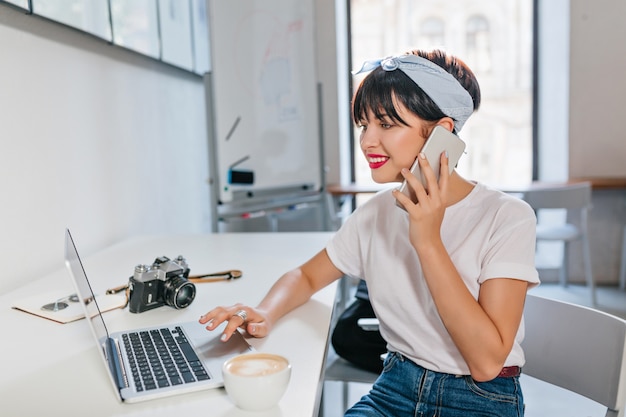 Lightly tanned smiling girl with dark shiny hair talking on phone while surfing in internet in her room