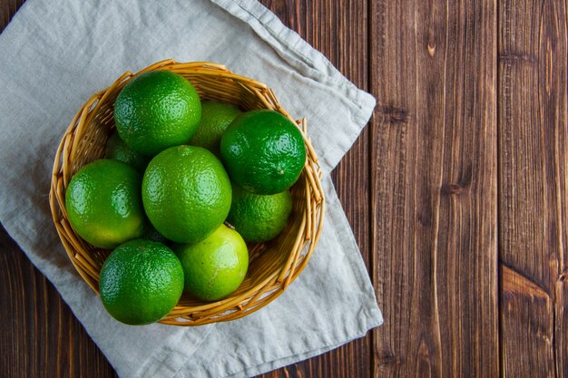 Limes in a wicker basket on wooden and kitchen towel, flat lay.