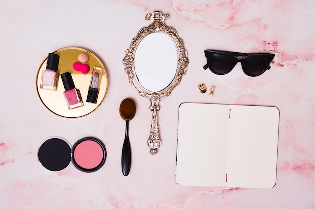 Lipstick; nail polish bottles; compact face powder; makeup brush; hand mirror; clutcher and open blank diary on pink backdrop