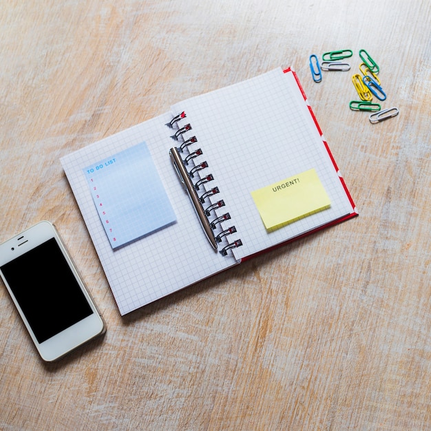 To do list notepad and urgent sticky note on checkered notebook with smartphone and paperclip