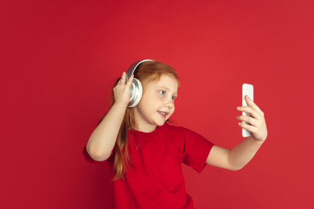 Listen to music. Caucasian little girl portrait isolated on red  wall. Cute redhair model in red shirt. Concept of human emotions, facial expression. Copyspace.