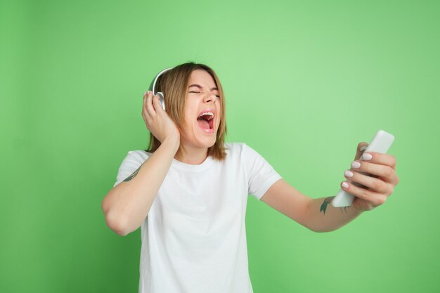 Listen to music, screaming. Caucasian young woman's portrait isolated on green  wall. Beautiful female model in white shirt. Concept of human emotions, facial expression, youth.