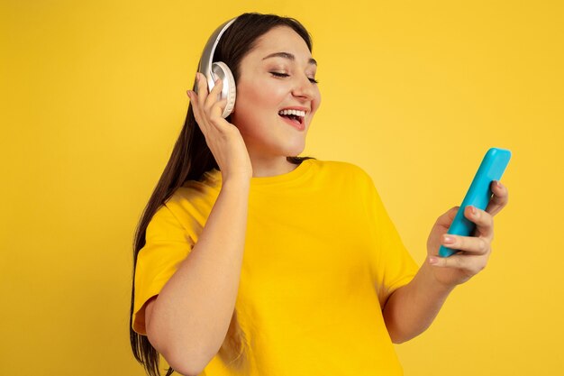 Listen to music with wireless headphones and phone. Caucasian woman on yellow studio background. Beautiful brunette model in casual. Concept of human emotions, facial expression, sales, ad, copyspace.