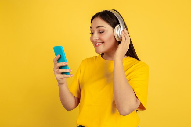 무선 헤드폰과 휴대폰으로 음악을 듣습니다. 노란색 벽에 백인 여자입니다. 캐주얼에 아름 다운 갈색 머리 모델입니다. 인간의 감정, 표정, 판매, 광고, copyspace의 개념.