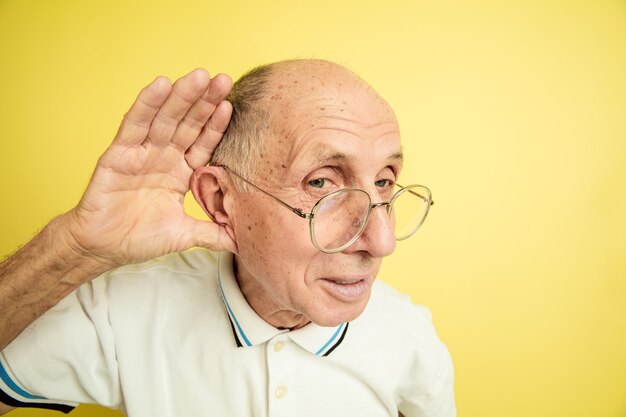 Listen to secrets. Caucasian senior man's portrait isolated on yellow studio background. Beautiful male emotional model. Concept of human emotions, facial expression, sales, wellbeing, ad. Copyspace.