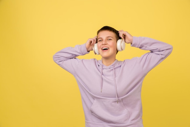 音楽を聴きます。コピースペースと黄色の背景に白人の女の子の肖像画。パーカー、ヘッドフォンの美しい女性モデル。人間の感情、顔の表情、販売、広告、ファッションの概念。