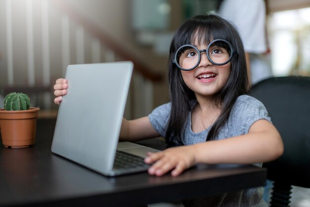 Маленькая очаровательная дочь в очках весело проводит время, играя на ноутбуке, притворяясь, что работает дома