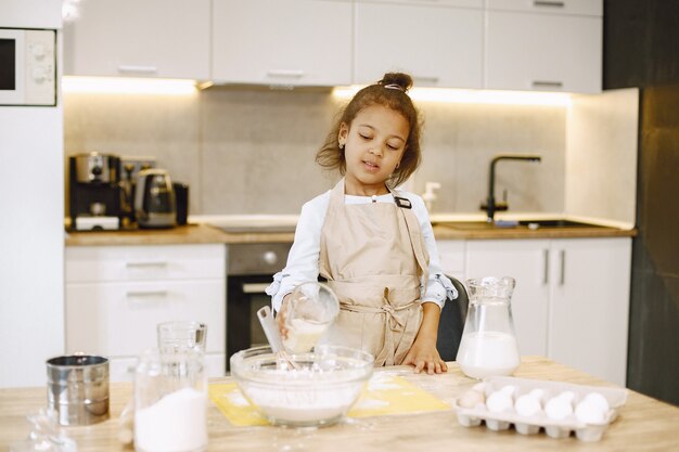 Little african-american girl pouring milk in a glass bowl, preparing a dough.