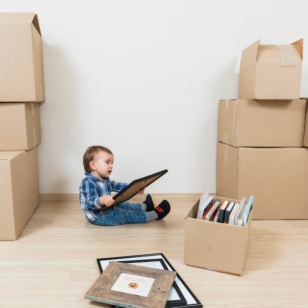 Little baby boy sitting between the moving cardboard boxes looking at picture frame at new home