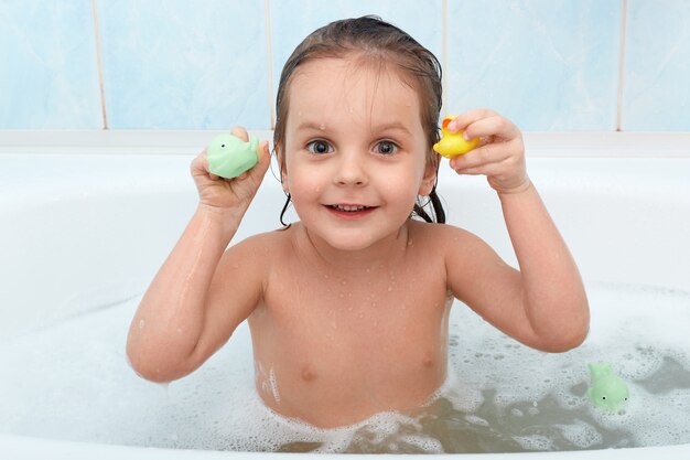 Little baby girl holding toy in hands taking bath, plays in water with duck and dolphin