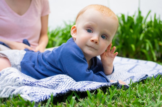 Piccola neonata sdraiata sul plaid, tenendo la mano vicino al viso e guardando lontano. closeup ritratto in giardino. giovane madre seduta. tempo di famiglia estivo, giornate di sole e concetto di aria fresca