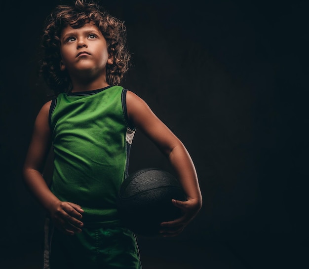 Little basketball player in sportswear holding ball in a studio. Isolated on a dark textured background.