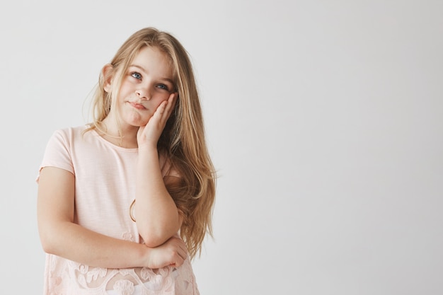 Little beautiful blonde girl with blue eyes squeezing face with hand, looking aside with frustrated expression after mom knows about bad marks at school.