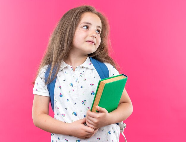 Little beautiful girl with long hair with backpack holding notebook looking aside with smile on happy face standing over pink wall