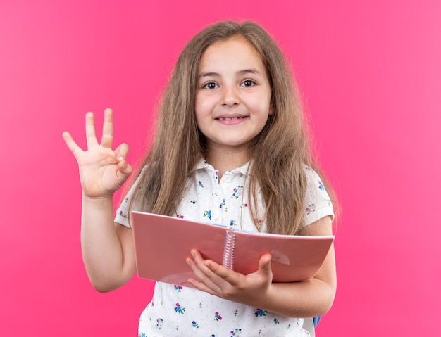 Free photo little beautiful girl with long hair with backpack holding notebook looking at front happy and positive showing ok sing smiling cheerfully standing over pink wall