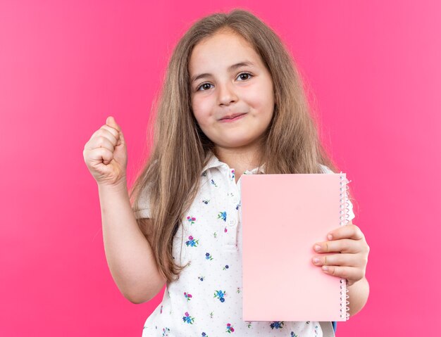 Little beautiful girl with long hair with backpack holding notebook looking at front with smile on happy face showing thumb up standing over pink wall