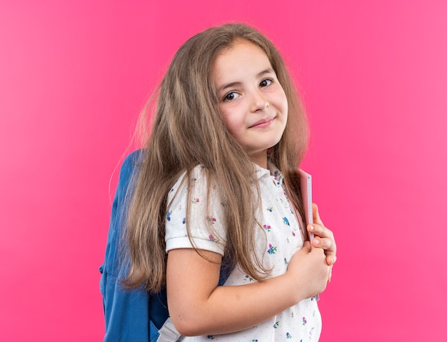 Little beautiful girl with long hair with backpack holding notebook looking at front with smile on happy face standing over pink wall