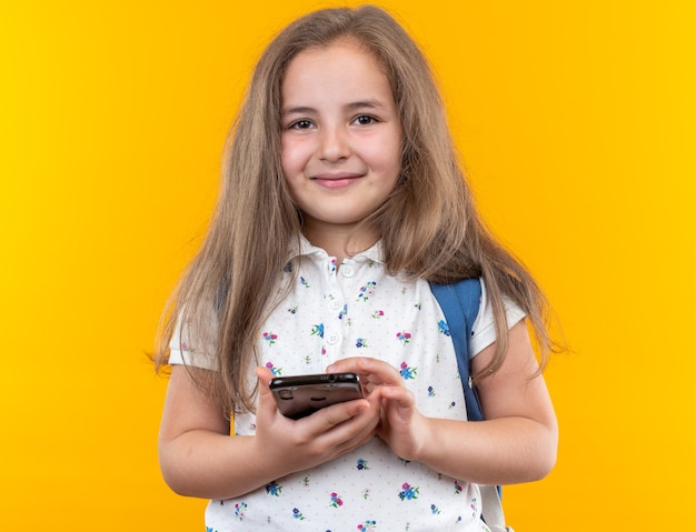 Маленькая красивая девушка с длинными волосами с рюкзаком держит смартфон счастливой и веселой улыбкой, стоя на оранжевом