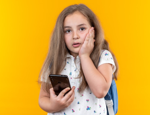 Little beautiful girl with long hair with backpack holding smartphone looking at front worried holding hand on her cheek standing over orange wall