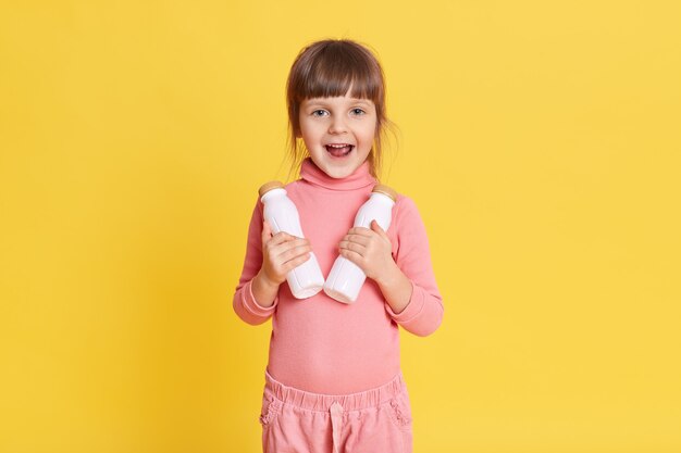 黄色のミルクの2本のボトルを保持している茶色の髪のピンクの服を着た小さな美女 無料の写真
