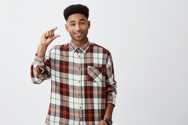 A little bit. Portrait of young good-looking dark-skinned funny guy with afro haircut in checkered shirt smiling, gesticulating with hands with sarcastic expression.