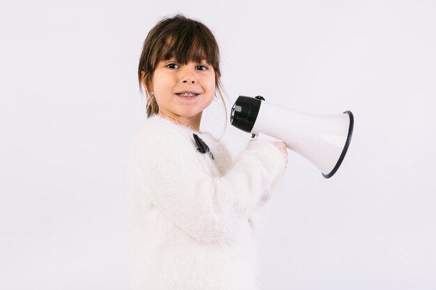 Маленькая черноволосая девочка в белом свитере с галстуком-бабочкой, говоря через белый мегафон.