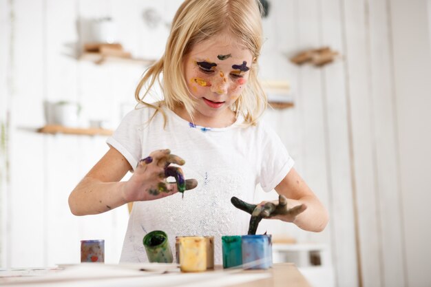 Little blond girl deeping her fingers in paint. European female child occupied with painting, wearing white t-shirt with paint spots on her face. Children and art.